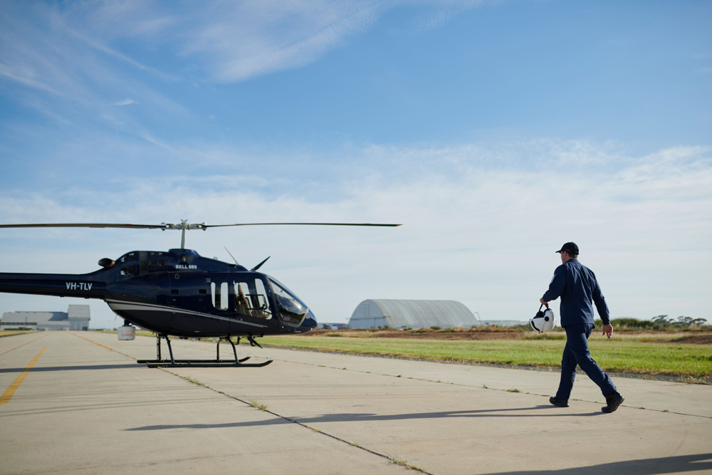 A man walking to a helicopter with an airfield and hangar in the background.